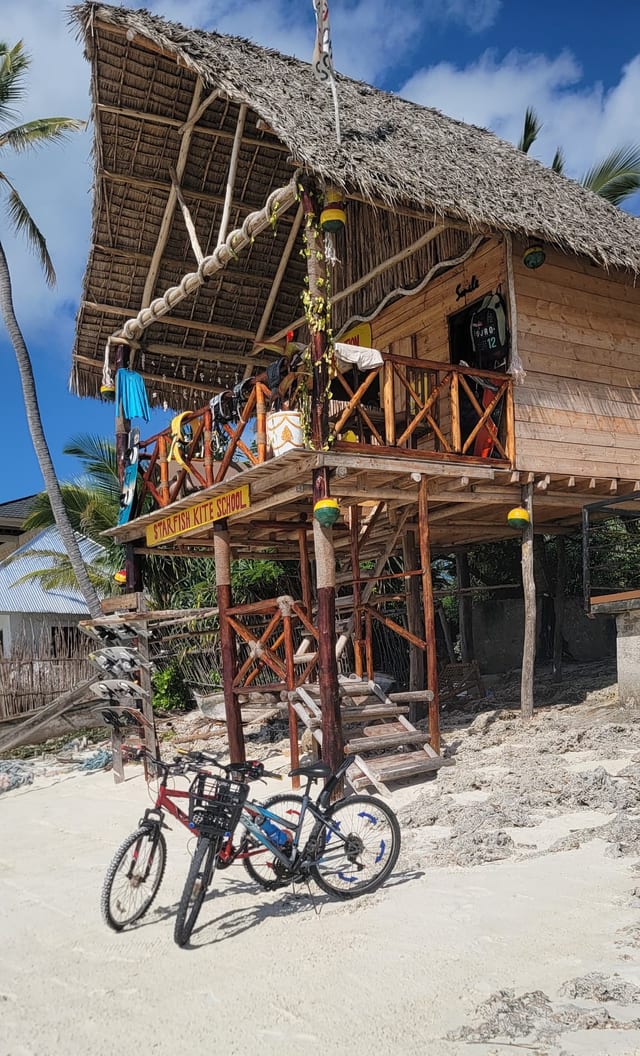 La capanna di Starfish Kite School su palafitte, due piani col tetto di makuti, l'insegna dipinta sopra la scaletta e due biciclette appoggiate sulla sabbia bianca.