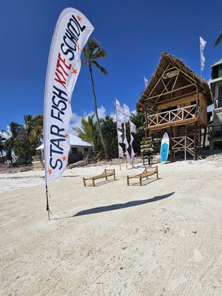 Una bandiera Starfish Kite School tesa nel vento, dietro la capanna e la rastrelliera delle tavole, i lettini di bambù sulla sabbia aperta.