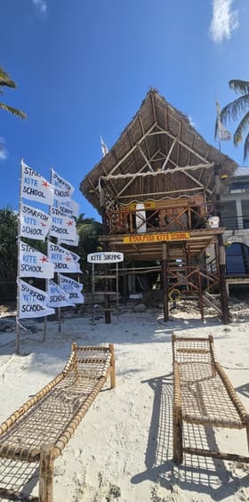 La capanna della scuola di kite vista di fronte, dietro una fila di bandiere Starfish, due lettini di bambù intrecciato sulla sabbia davanti.