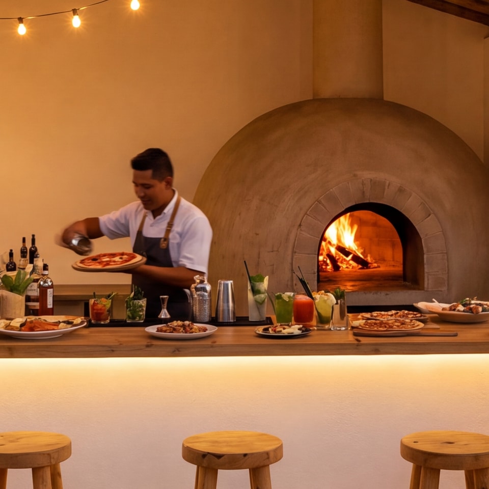 Il bar in servizio: il cuoco porta una pizza accanto al forno a cupola acceso, i bicchieri e i piatti lungo il bancone, le lucine sopra.